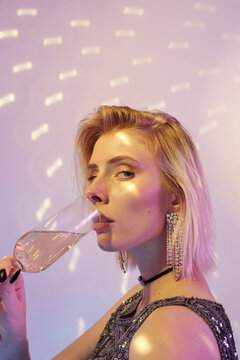 Portrait of stylish girl standing in lights drinking champagne at party looking at camera