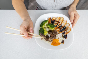 A girl in a gray shirt in the kitchen at home holds a plate of golden rice, shiitake mushrooms, chicken with unagi sauce and broccoli in her hands. Asian food concept