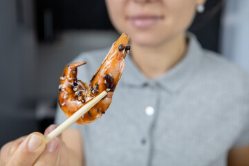 A girl in a gray shirt in the kitchen holds a king prawn in unagi sauce and sesame seeds with chopsticks. Asian food concept
