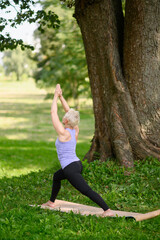 Middle aged woman doing yoga outdoors in warrior pose, Virabhadrasana I. Sports concept in city park.