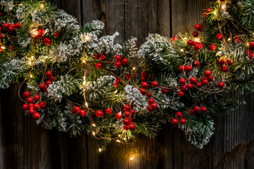 Fototapeta na wymiar Traditional Christmas wreath with red berries and sparkling garland. Cozy winter rustic background with Christmas wreath on a wooden wall. 