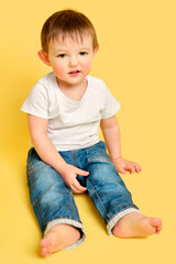 Portrait of an embarrassed toddler baby in full length on a studio yellow background. The child is sitting on the floor in a white T-shirt and blue jeans. Kid aged one year and four months