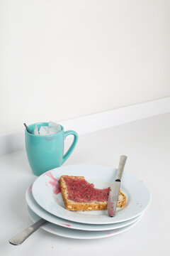 Restos de tostada con mermelada sobre montaña de platos sucios y taza de desayuno en fondo blanco vertical