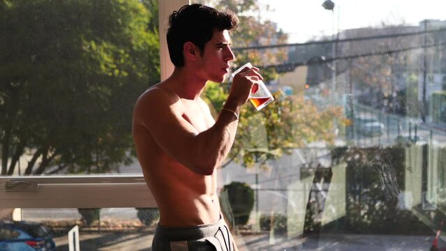 Sexy young man standing shirtless by curtains with soda or tea
