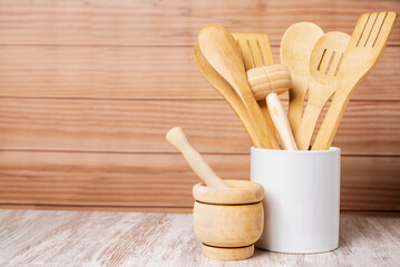 Wooden kitchen utensils inside a jar and next to a mortar, spoon, fork, colander and tongs among others, arranged in a decorative way with a light wooden background, copy space,