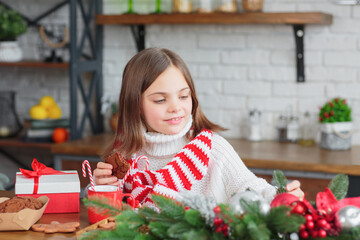 Cute smiling little child girl eating chocolate biscuits and drinking hot cocoa in mug sitting in a christmas decorated kitchen. Kid wear white knitted sweater. Xmas holidays