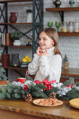 Cute smiling little child girl eating chocolate biscuits and drinking hot cocoa in mug sitting in a christmas decorated kitchen. Kid wear white knitted sweater. Xmas holidays