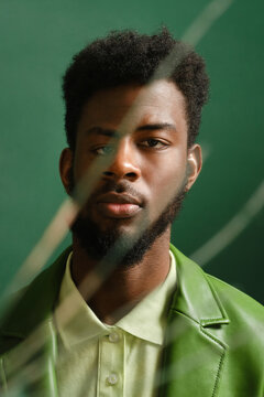 Unusual portrait shot of handsome young Black man wearing stylish green leather jacket and shirt standing against green wall background looking at camera