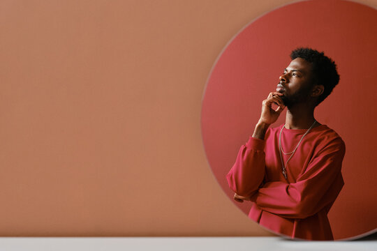 Vertical creative mirror reflection studio portrait shot of stylish young African American man wearing coral pink sweatshirt thinking of something, copy space