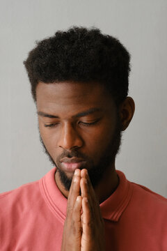 Vertical close-up studio portrait of modern young African American man with beard on face wearing pink T-shirt posing on camera with praying hands gesture, gray background