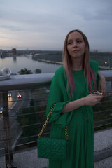 Obraz premium Vertical portrait of a beautiful stylish woman enjoying evening out in the city, standing on a glass bridge over busy car road