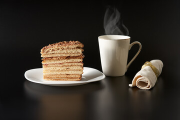 A piece of cake made of many thin cakes held together by oustard on a white plate on a black background. Next to the white cup and napkin.