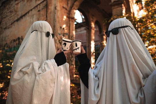 A funny image of two people in ghost costumes and sunglasses holding a cup with a drink in an abandoned building