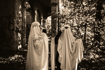 Naklejka premium A funny image of two people in ghost costumes and sunglasses holding a cup with a drink in an abandoned building, black and white image