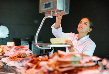 Interested young saleswoman working at counter in supermarket, weighing piece of semi-hard cheese on scales ..