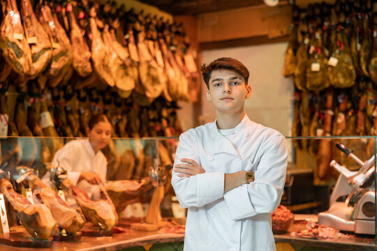 Confident young guy, butcher shop salesman in white uniform, standing with crossed arms on background of counter with hanging jamon legs