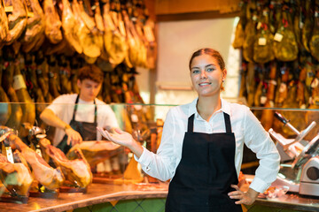 Successful smiling young woman, butcher shop owner, standing in apron near glass counter, greeting...