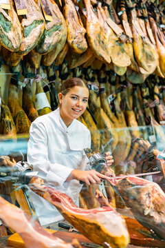 Cheerful young female seller in white uniform cutting slices from whole leg of dry-cured Iberian jamon on jamonera in butcher store