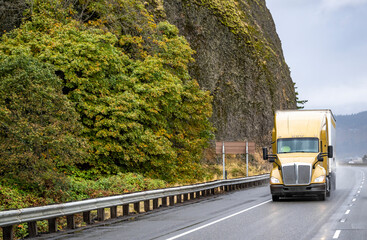 Fototapeta premium Gold big rig semi truck transporting cargo in refrigerator semi trailer driving on the wet mountain road with rock cliff on the side at raining weather