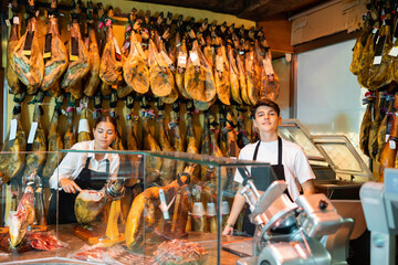 Fototapeta na wymiar Skilled young salesgirl and salesman of butcher shop selling delectable dried Iberian jamon, working together at counter