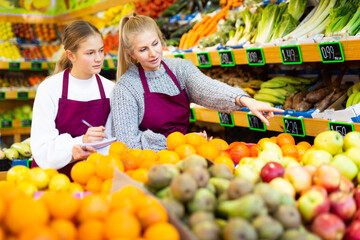 Obraz premium Young trainee salesgirl at greengrocery store listening attentively to experienced female colleague and making notes. First days of work concept