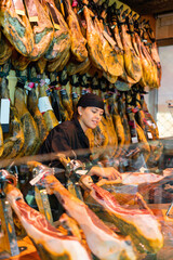 Fototapeta na wymiar Attentive female seller cutting slices from whole leg of jamon in meat store