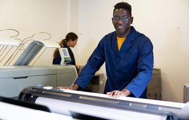 Positive middle-aged African American man in uniform using plotter during work in the printing...