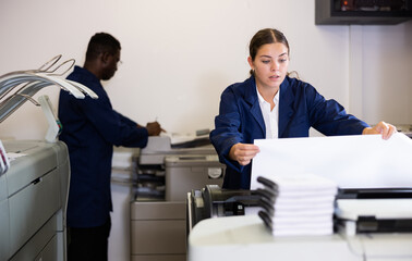 Serious young woman in uniform loading large format paper in a plotter in the print shop