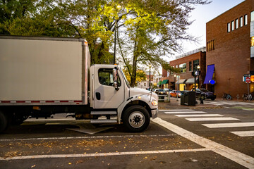 Local day cab white big rig semi truck with box trailer standing on the crossroad on evening small city street waiting for the green traffic light