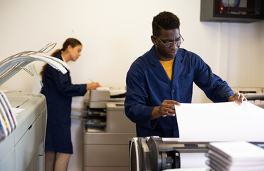 Serious middle-aged African American man in uniform loading large format paper in a plotter in the...