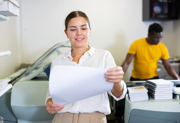 Fototapeta premium Attentive young female specialist verifying printed sheet during work in the print shop