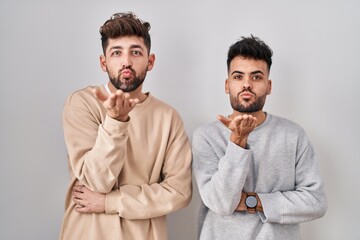 Young homosexual couple standing over white background looking at the camera blowing a kiss with hand on air being lovely and sexy. love expression.