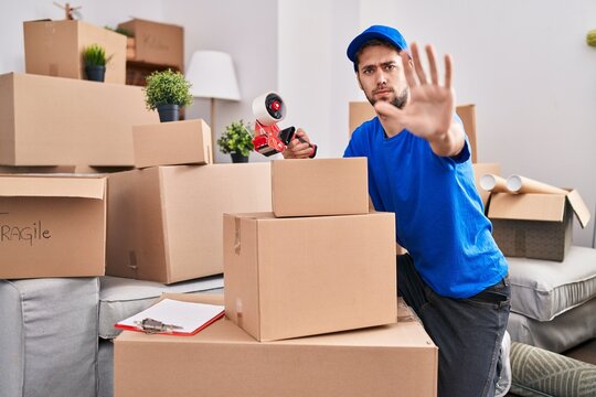 Hispanic man with beard working moving boxes with open hand doing stop sign with serious and confident expression, defense gesture