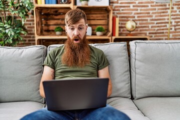 Redhead man with long beard using laptop sitting on the sofa at the living room scared and amazed...