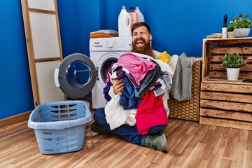 Redhead man with long beard putting dirty laundry into washing machine winking looking at the...
