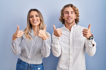 Young couple standing over blue background success sign doing positive gesture with hand, thumbs up smiling and happy. cheerful expression and winner gesture.