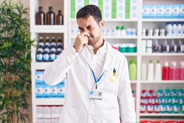 Handsome hispanic man working at pharmacy drugstore smelling something stinky and disgusting,...