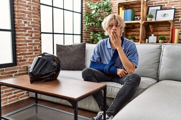 Young man wearing arm on sling for motorcycle accident covering mouth with hand, shocked and afraid...