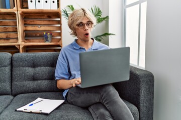 Young man working using computer laptop sitting on the sofa scared and amazed with open mouth for surprise, disbelief face