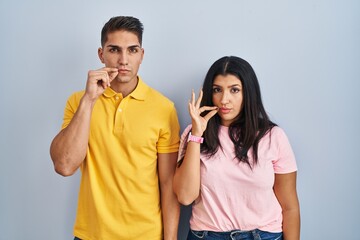 Young couple standing over isolated background mouth and lips shut as zip with fingers. secret and...
