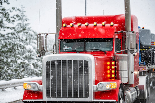 Stylish bright red classic big rig semi truck with turned on auxiliary marker lights transporting cargo on flat bed semi trailer slowly skid on a winter snowy highway road