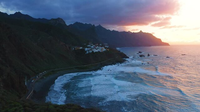 Small village on mountainside by ocean at dark cloudy night. Aerial overview of sunset light cover rural town surrounded by sea waters. Almaciga Tenerife Canary Island Spain.