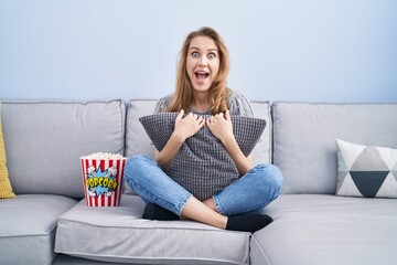 Obraz premium Beautiful blonde woman watching tv sitting on the sofa celebrating crazy and amazed for success with open eyes screaming excited.