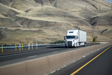 White color big rig high cab long haul semi truck transporting frozen cargo in refrigerated semi trailer running on divided highway road with bald mountains on background