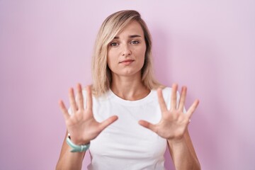 Fototapeta premium Young blonde woman standing over pink background moving away hands palms showing refusal and denial with afraid and disgusting expression. stop and forbidden.