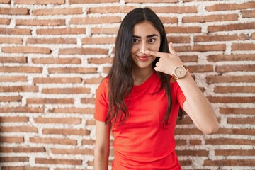 Young teenager girl standing over bricks wall pointing with hand finger to face and nose, smiling cheerful. beauty concept
