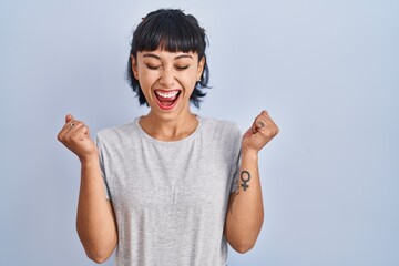 Young hispanic woman wearing casual t shirt over blue background excited for success with arms...