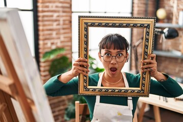 Young beautiful woman sitting at art studio with empty frame in shock face, looking skeptical and...