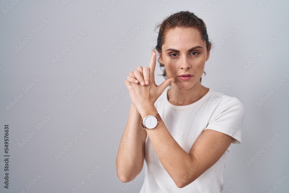 Sticker Beautiful brunette woman standing over isolated background holding symbolic gun with hand gesture, playing killing shooting weapons, angry face