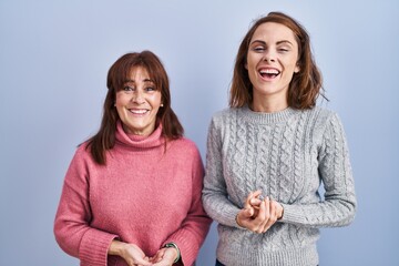 Mother and daughter standing over blue background with hands together and crossed fingers smiling...